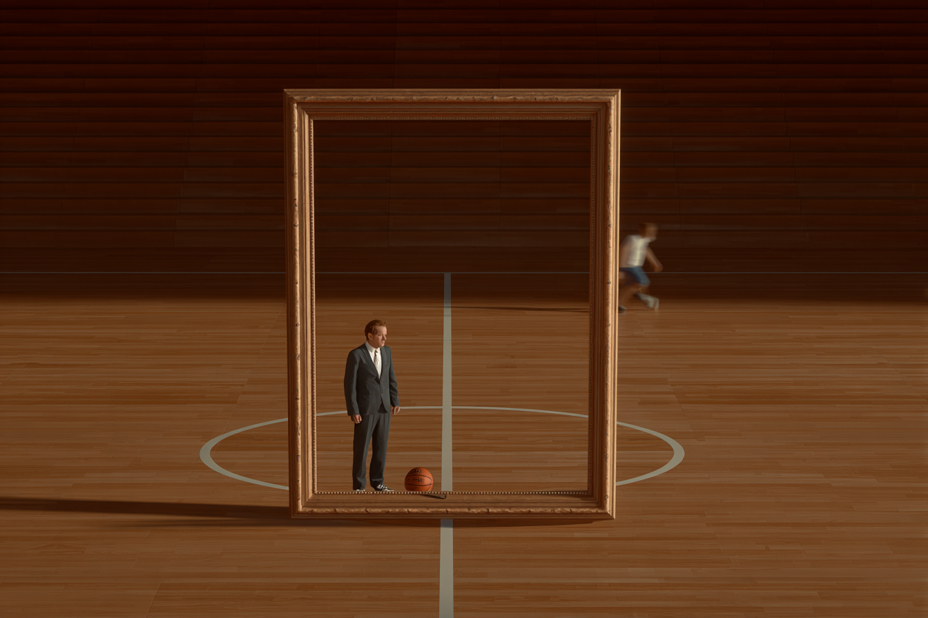 A man in a suit stands inside an empty picture frame on a basketball court while someone is playing basketball.