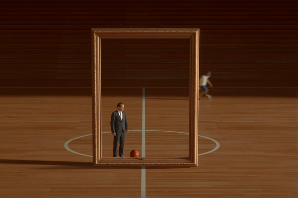 A man in a suit stands inside an empty picture frame on a basketball court while someone is playing basketball.